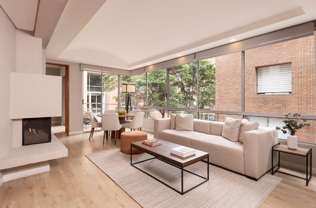Sala de un apartamento en Bogotá con ventanales de piso a techo, sofá en lino claro, mesa de centro en madera y chimenea blanca