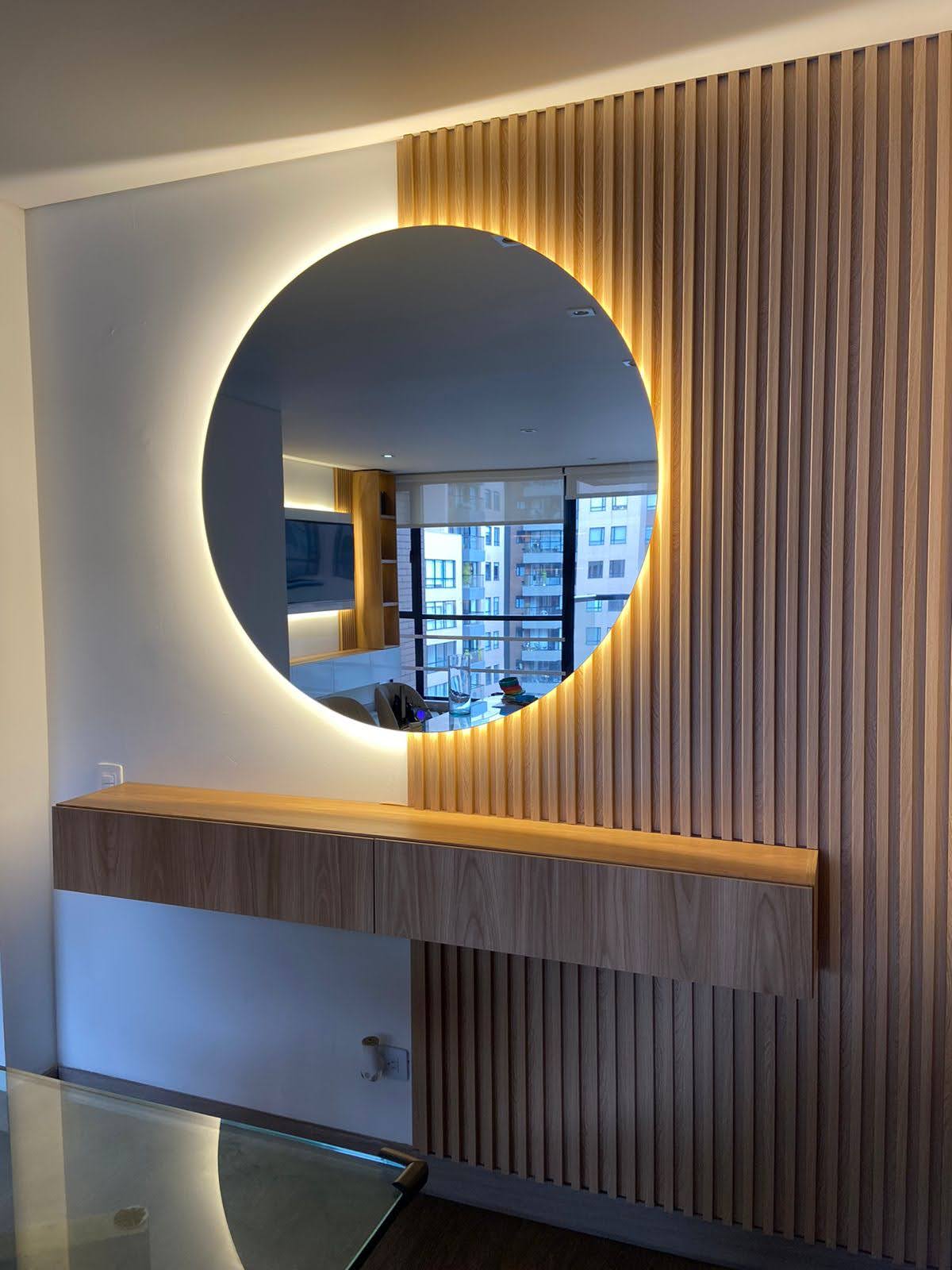 Espejo circular retroiluminado sobre consola flotante y panel de listones de madera